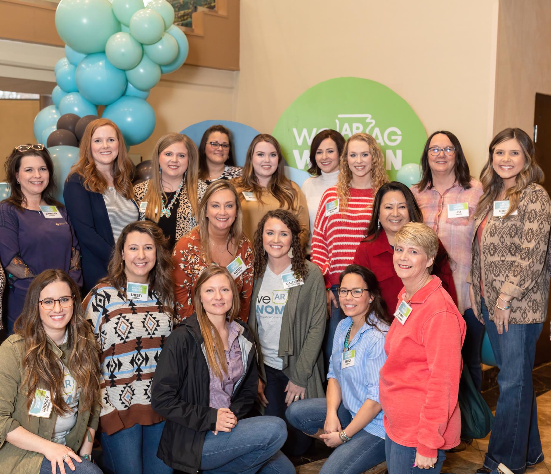 16 smiling ladies standing in front of teal balloons at the AR Women in Ag conference.