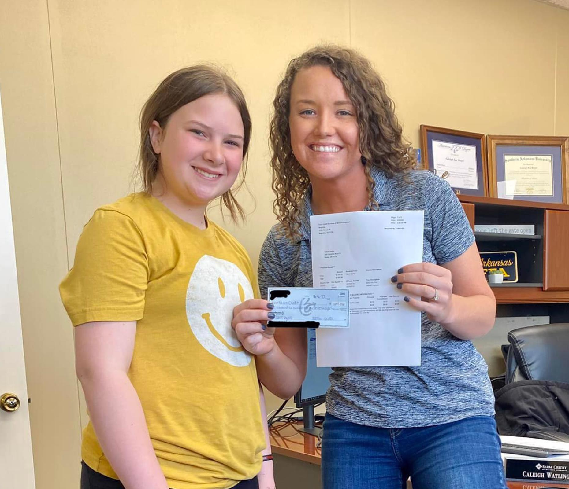 young girl in yellow shirt with happy face on it smiles as Loan Officer Caleigh Watlington shows her paid off youth loan paperwork.