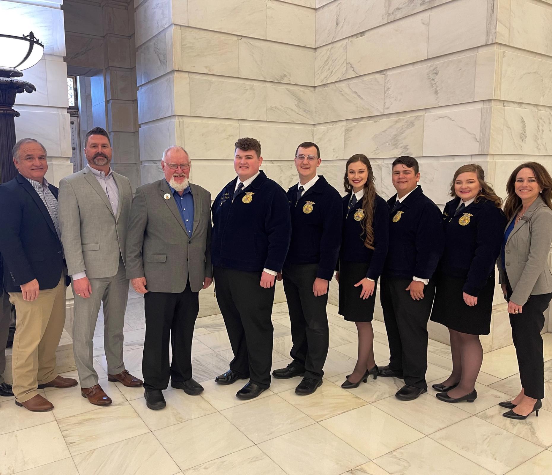 6 people standing in front of a white marble wall at the AR State Capitol. Four on the right are wearing official FFA jackets and photo recognizes Farm Credit's $50,000 donation to updating Camp Couchdale.