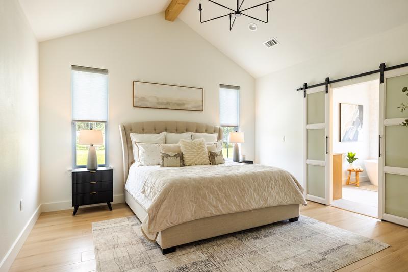Greene family bedroom with large beige tufted headboard and neutral color bedding. Side tables with lamps, barn doors leading to master bathroom.