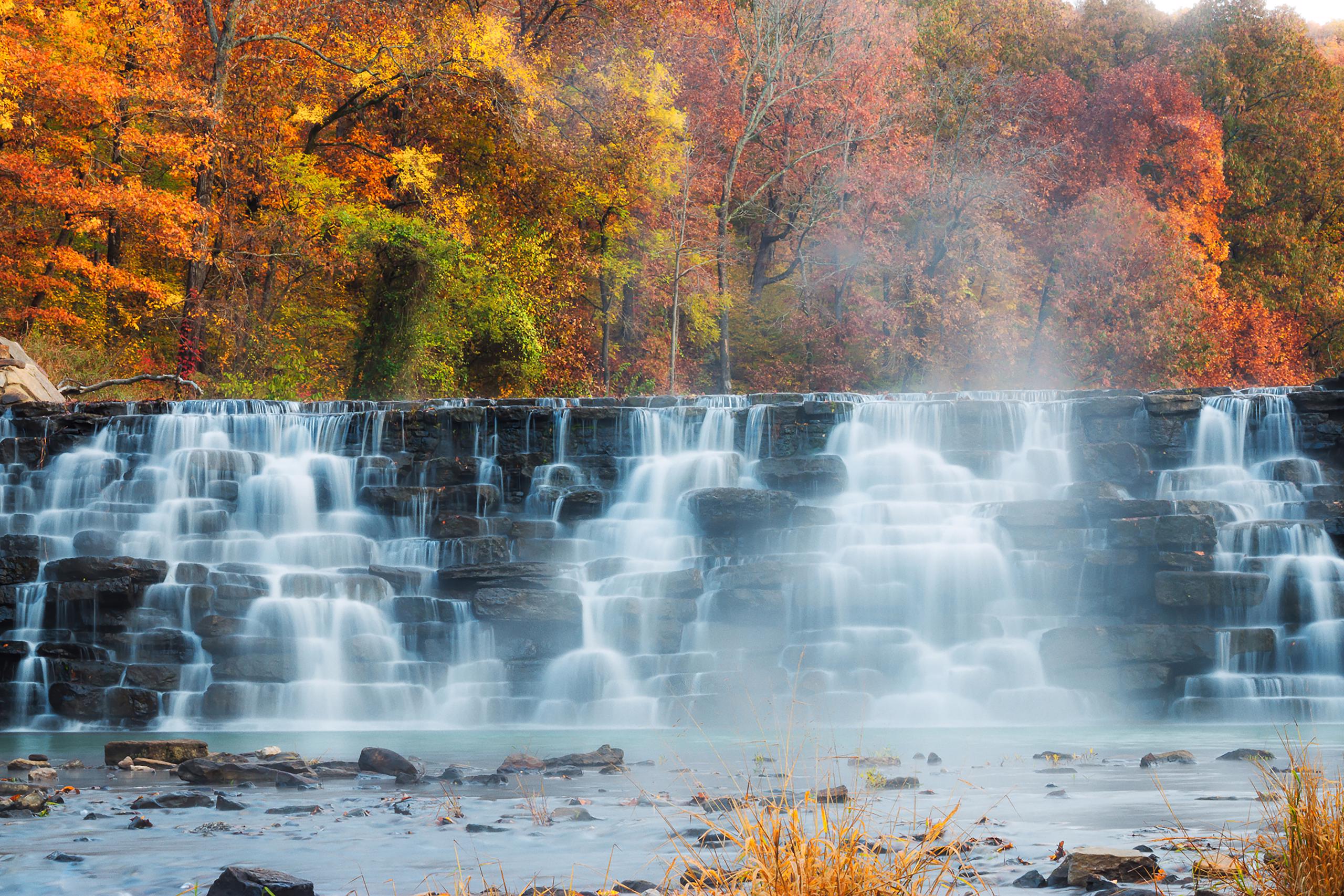 A misty rock wall waterfall in front of vibrant orange, yellow, and green trees.