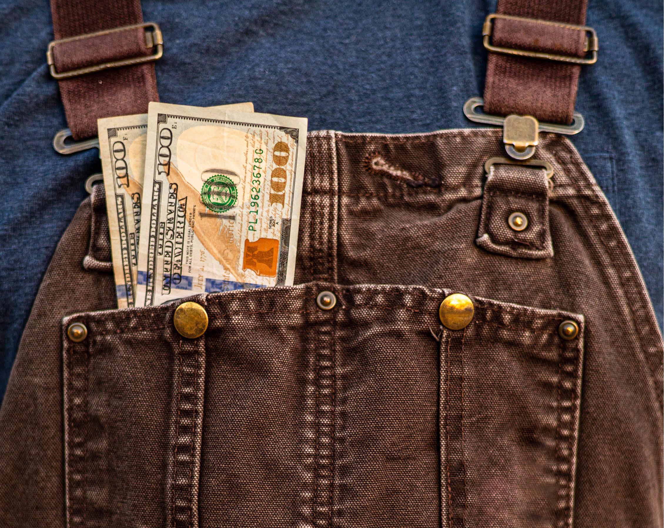 Two $100 bills stick out of pocket of brown overalls.