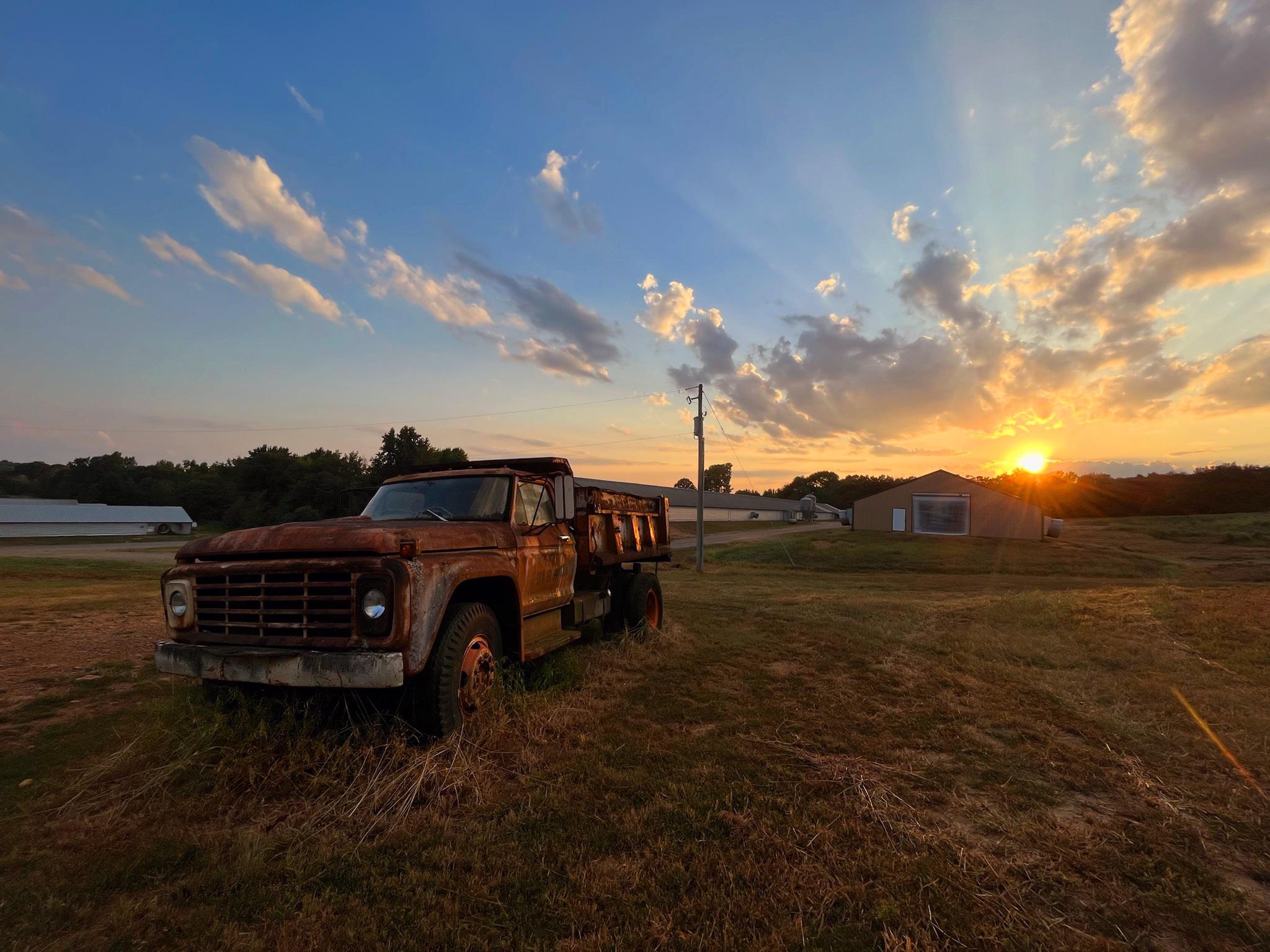 Old rusty truck in foreground, chicken houses in middle ground, sunset sky.